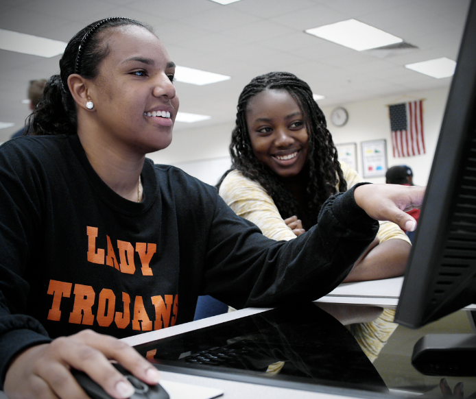 Two students are sitting together at a computer workstation in a classroom. The student on the left is wearing a black "Lady Trojans" sweatshirt and is using the computer mouse, while the student on the right is smiling and pointing at the computer screen. An American flag and other classroom decorations are visible in the background. The atmosphere appears collaborative and positive.